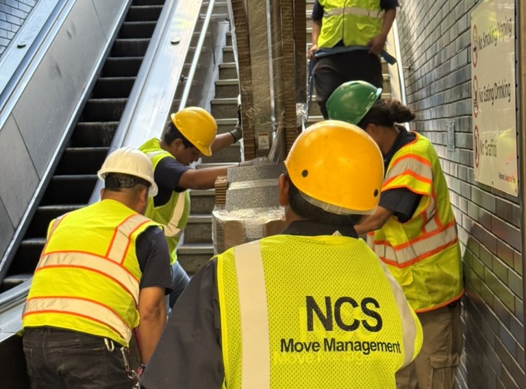 NCS crew carrying wrapped equipment through a transit station stairwell