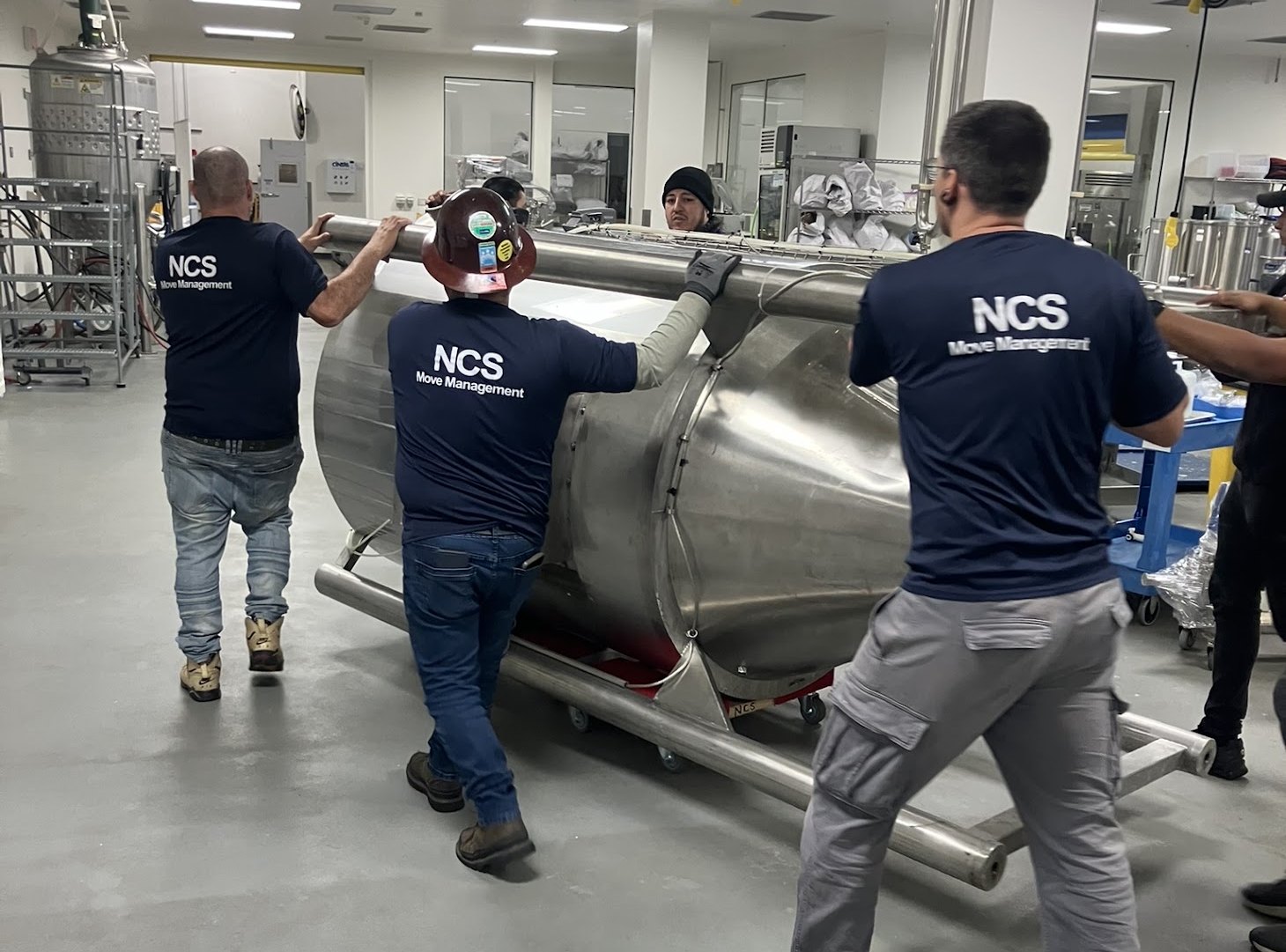 NCS crew moving a stainless fermenter vessel on skates through a cleanroom